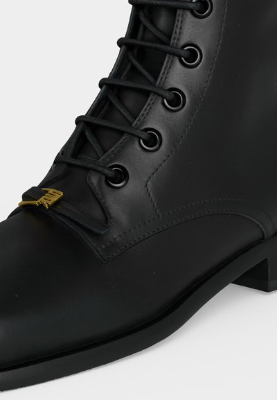 Bottine en cuir noir avec des lacets noirs passés dans des œillets métalliques et un détail métallique doré au-dessus des orteils.