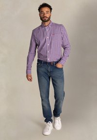 Geruite paarse en witte shirt, blauwe jeans en witte sneakers. Het shirt heeft een button-down kraag en een zak; de jeans zijn recht gesneden.