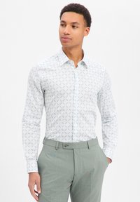 Camicia bianca con bottoni e motivo floreale verde chiaro, maniche lunghe e colletto appuntito. Abbinata a pantaloni sartoriali grigio chiaro.