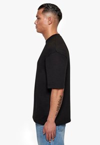 Black cotton t-shirt with short sleeves and a crew neck, featuring a relaxed fit and a smooth texture. Worn with light blue jeans.