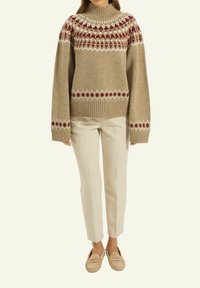 Beige gestrickter Pullover mit hohem Kragen, der lebhafte Muster in Kastanienbraun und Weiß über der Brust und an den Ärmeln aufweist. Kombiniert mit hellen Hosen.