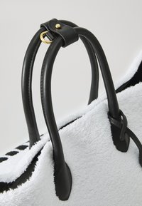 Čierne kožené rúčky so zlatými kovovými detailmi pripevnené na mäkkej, bielej taške s textúrou, ktorá má čierne akcenty a charakteristický tvar.