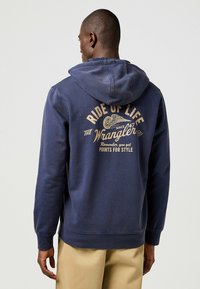 Marinblå hoodie med en känguruficka, prydd med stor guldfärgad text på ryggen med orden "RIDE OF LIFE" och "THE Wrangler."