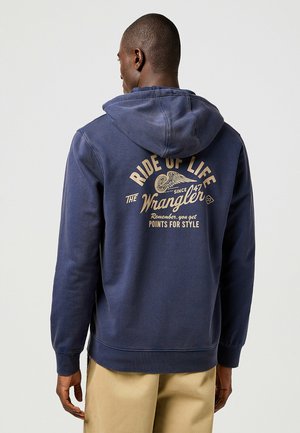 Navyblauer Kapuzenpulli mit Kordelzugkapuze, ausgestattet mit einem großen grafischen Print auf der Rückseite in beigen Text- und Designelementen. Material aus Baumwollmischung.