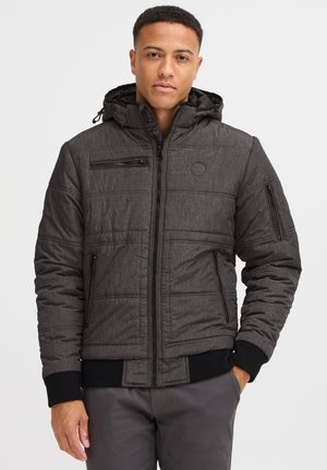 Veste noire matelassée avec capuche, fermeture éclair à l'avant et poignets côtelés ; elle possède des poches zippées sur la poitrine et sur les côtés, un tissu texturé et un logo en patch.
