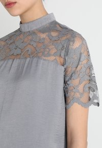 Top gris con un escote alto, mangas cortas con intrincados detalles de encaje gris y un cuerpo de tela suave y ligeramente texturizada.