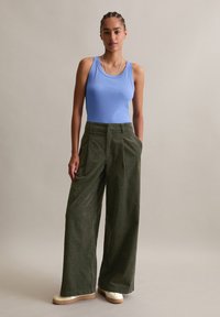 Hellblaues Tanktop kombiniert mit olivgrünen weit geschnittenen Hosen. Der matte Stoff hat vordere Falten und seitliche Taschen, ergänzt durch weiße Sneakers.