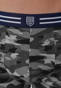 Záber zblízka osoby nosiacej šedé, čierne a biele spodné prádlo s kamuflážnym vzorom, s námorníckym pásom s bielymi pruhmi a emblémom.