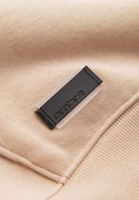 Beiger Stoff mit einem schwarzen und klaren rechteckigen Kleidungslabel, das mit dem Markennamen „ombre“ geprägt ist, nahe einer Naht eingenäht.