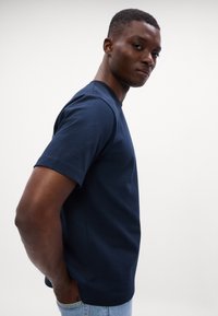 Tamsiai mėlyna medvilninė marškinėliai su apykakle ir trumpomis rankovėmis, pasižymintys laisvu siluetu ir glotnia tekstūra. Paprastas dizainas be raštų.