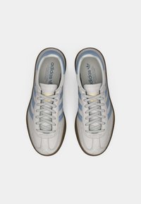 Sapatilhas Adidas em cinza claro com detalhes em azul, apresentando atacadores planos, uma sola de borracha texturizada e marcação na lingueta e na palmilha.