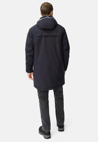 Mørkeblå vandtæt parka med hætte, lang ryg og justerbare manchetter, parret med mørke jeans og sorte robuste støvler.