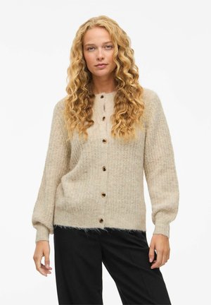 Mulher com cabelos longos e encaracolados, loiros, vestindo um cardigan de malha bege com botões e calças pretas, de pé contra um fundo branco simples.
