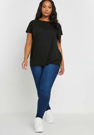 Vrouw met lang zwart haar, gekleed in een zwart t-shirt, blauwe skinny jeans en witte platformsneakers, die naar voren loopt tegen een effen achtergrond.