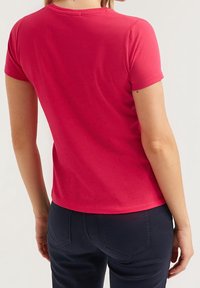 Žena v jednoduchém červeném tričku s krátkým rukávem a tmavě modrých těsných kalhotách, zobrazená zezadu na neutrálním pozadí.