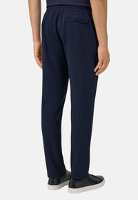 Tamsiai mėlynos elastingos liemens kelnės su užpakaline kišene, siaurėjančio kirpimo ir lygia tekstūra. Dėvimos su juodais sportbačiais ant baltos fono.