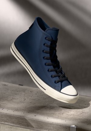 Baskets montantes avec une tige en toile texturée bleu marine, un capuchon de protection en caoutchouc blanc, des lacets noirs et deux rayures blanches sur la semelle.
