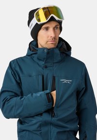 Teal waterdichte skijack met zwarte accenten, ritszakken, hoge kraag en verstelbare manchetten. Draag het met een zwarte beanie en skibril.