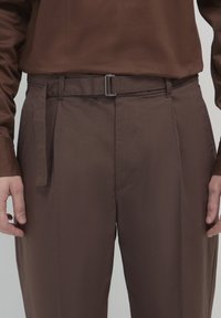Braune Hose aus glattem Stoff, ausgestattet mit einem Gürtel mit einer silbernen Schnalle, Seitentaschen und einem geraden Bein.