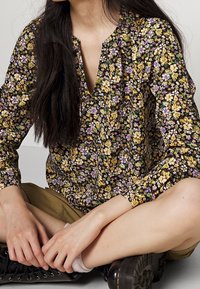 Sort blomstret bluse med små gule, lilla og hvide blomster; letvægtsstof; V-hals og lange ærmer; løs pasform.
