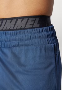 Zili sporta šorti ar teksturētu audumu, ar melnu elastīgu jostu, kurā pelēkā krāsā ir izdrukāts "HUMMEL". Minimalistisks dizains, viegls materiāls.