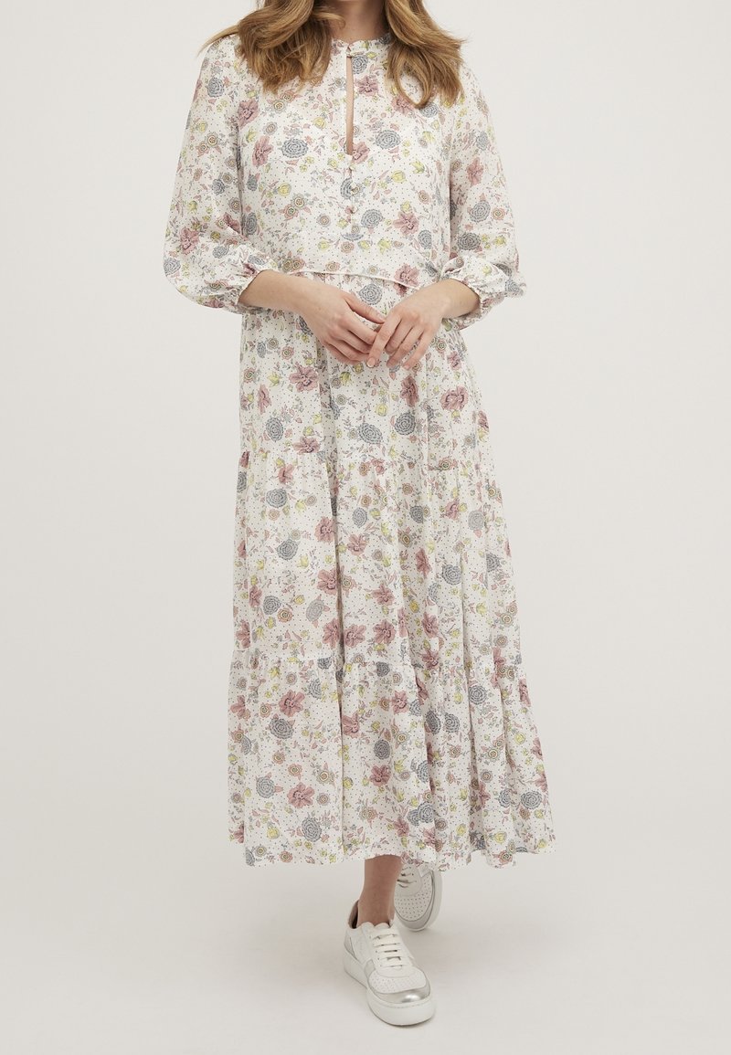 Femme portant une longue robe blanche à motifs floraux roses, jaunes et gris, assortie à des baskets blanches, debout sur un fond uni.