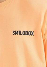Peachfarvet bomuld t-shirt med et sort trykt logo "SMILODOX" foran. Korte ærmer og rund halsudskæring. Blød tekstur.