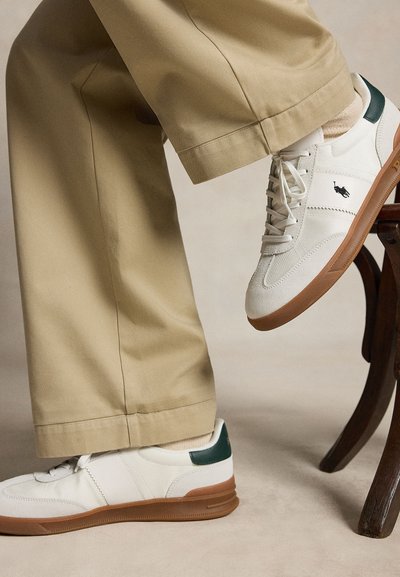 Osoba oblečená v béžových nohaviciach a bielych teniskách s hnedou podrážkou a tmavozeleným detailom na päte, sediaca s jednou nohou položenou na drevenej nohe stoličky.