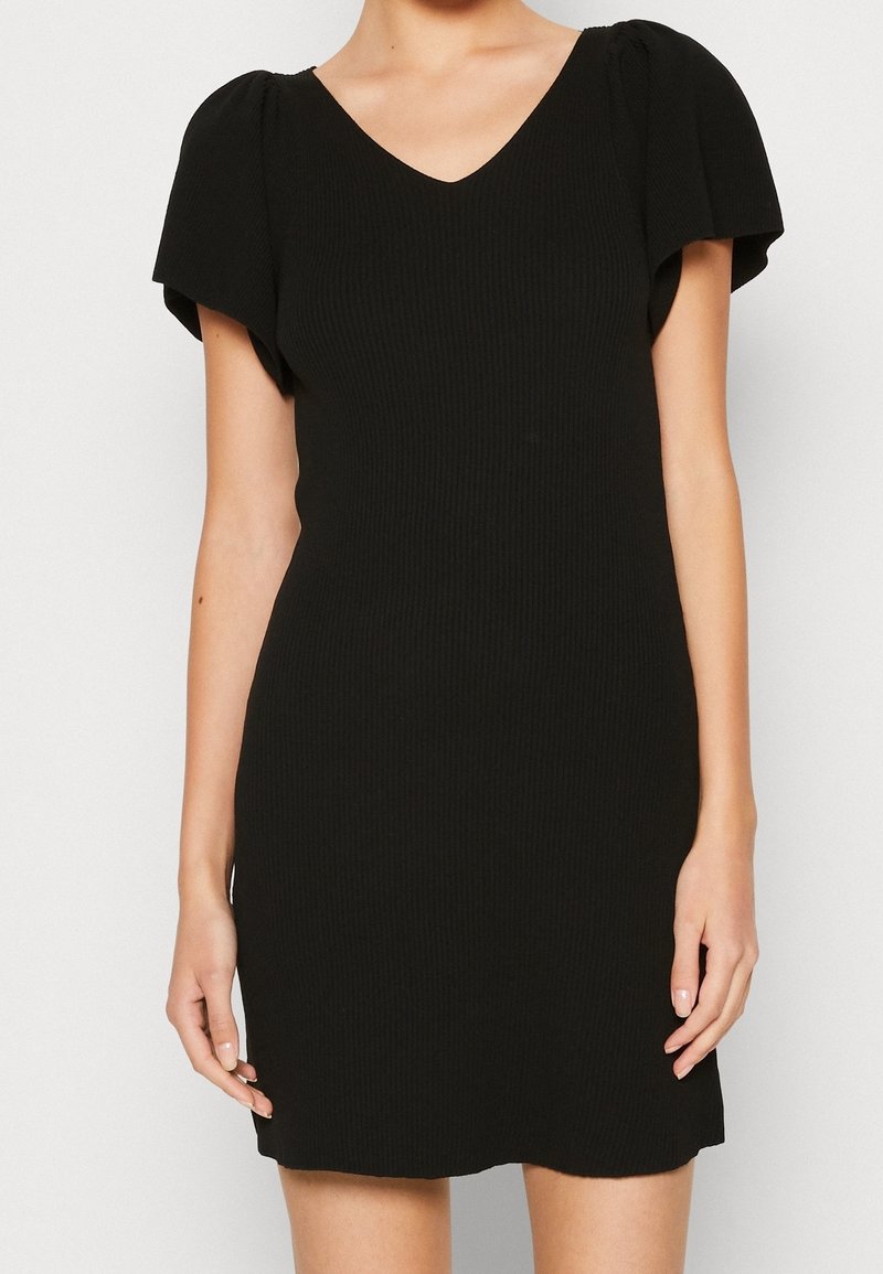 Femme portant une robe noire courte avec des manches courtes bouffantes et un décolleté en V, debout sur un fond blanc uni.