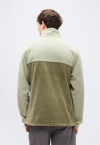 Φλις μπουφάν με ψηλό γιακά, διαθέτει διχρωμία σε ανοιχτό πράσινο και ελιά, υφή υφάσματος και ρυθμιζόμενο στρίφωμα.