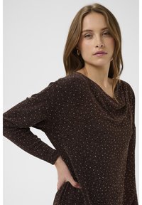 Pruun pika varrukaga topp, millel on drapeeritud kaelus ja valgete täppidega tekstuuritud pind. Sile kangas ja keha järgi istuv siluett.