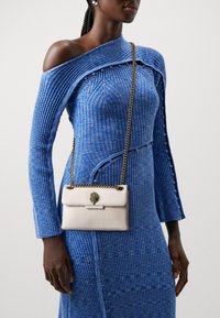 Borsa a tracolla bianca con texture e catena dorata, dotata di una chiusura decorativa, abbinata a un vestito blu a coste con taglio sulle spalle e bottoni.