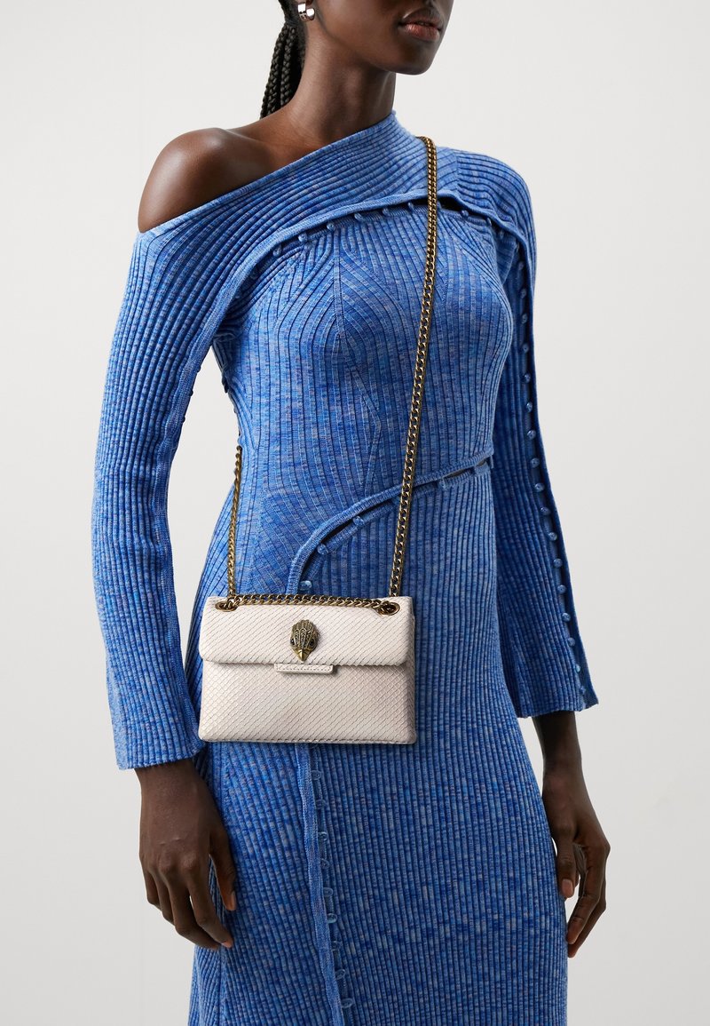 Borsa a tracolla bianca con texture e catena dorata, dotata di una chiusura decorativa, abbinata a un vestito blu a coste con taglio sulle spalle e bottoni.