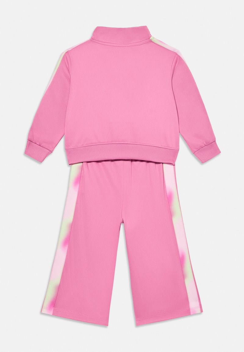 Completo sportivo rosa con giacca zip-up e colletto alto e pantaloni coordinati. La giacca presenta strisce a contrasto sulle spalle, mentre i pantaloni hanno dettagli laterali.