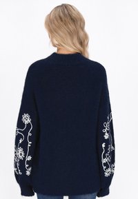 Marineblauwe gebreide trui met oversized mouwen en witte bloemenborduursels langs de armen. Zachte textuur en een ontspannen pasvorm.