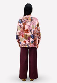 Frau mit langen schwarzen Haaren steht mit dem Rücken zur Kamera, trägt ein lockeres pinkes Shirt mit bunten Möbelmotiven und weiten burgunderroten Hosen.