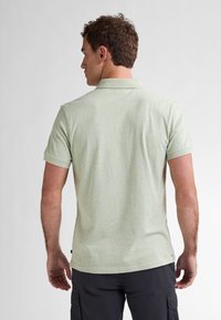 Polo shirt verde chiaro con texture a macchie, maniche corte e colletto. Esibito da dietro, abbinato a pantaloni cargo scuri.