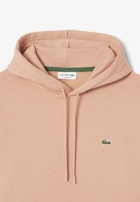 Sweat à capuche marron clair avec cordons, avec un petit logo crocodile vert sur la poitrine gauche et une étiquette "Lacoste Classic Fit" à l'intérieur du col.