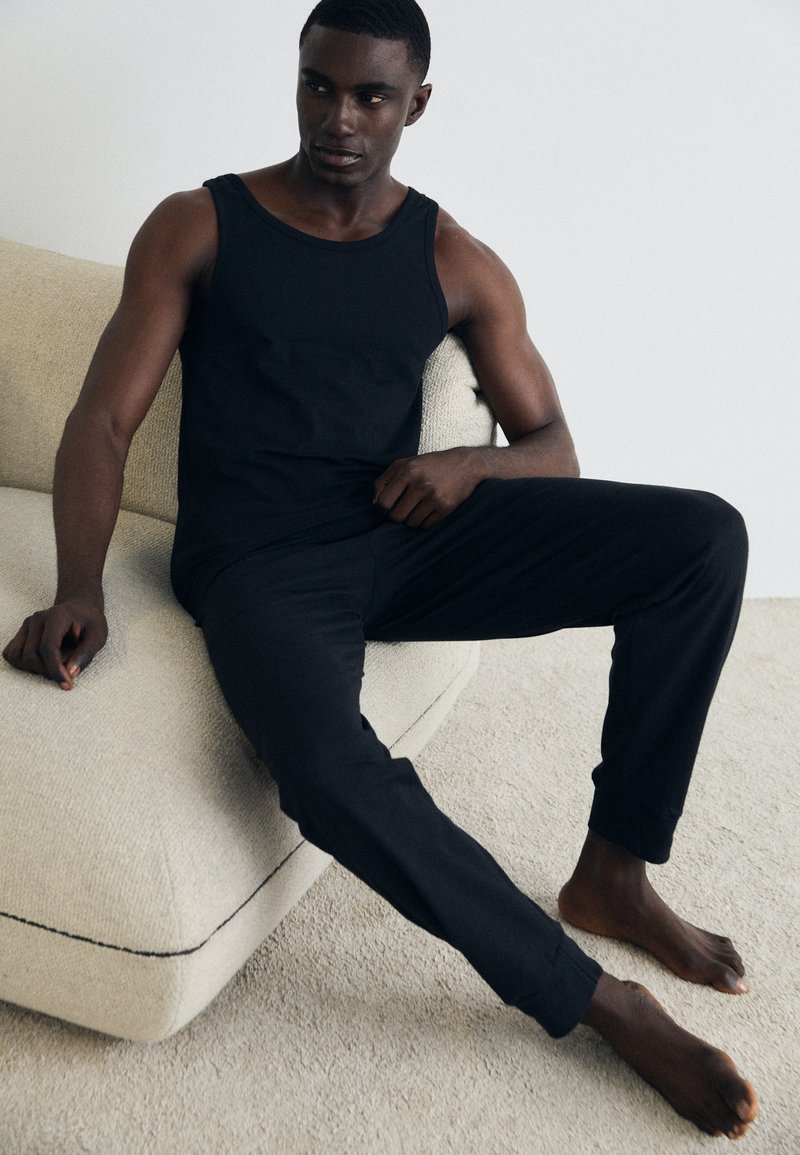 Sort tanktop og bukser lavet af blødt stof, tætsiddende design, glat tekstur og minimale sømme, båret af en siddende person på en lys sofa.
