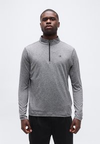 Νεαρός άνδρας με γκρι ζακέτα με φερμουάρ και μακριά μανίκια με διακριτικές ρίγες και μαύρο παντελόνι, στέκεται μπροστά σε λευκό απλό φόντο.