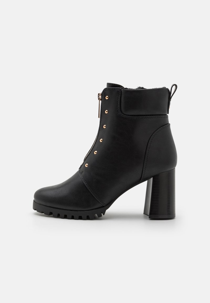 Bottine noire à cheville avec talon bloc, fermeture éclair frontale, bout rond, clous dorés et semelle robuste sur fond blanc.