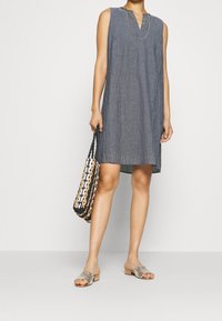 Robe droite grise sans manches en tissu texturé, avec un décolleté en V entaillé et des poches latérales, associée à un sac tressé et des sandales perlées.