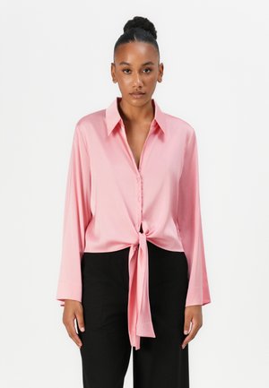 Rosa Satinbluse mit langen Ärmeln und einem gebundenen Knoten an der Taille. Sie verfügt über einen klassischen Kragen und eine durchgeknöpfte Vorderseite. Wird mit schwarzen Hosen kombiniert.