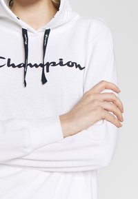 Felpa bianca con logo "Champion" in blu navy. Presenta un cappuccio con cordino e un tessuto morbido. I polsi sono a costine, evidenziando una vestibilità comoda.