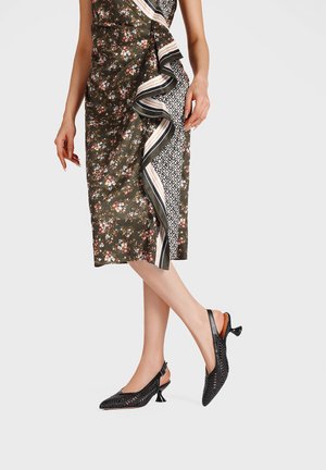 Donna con gonna a portafoglio floreale con dettaglio di volant e scarpe nere intrecciate slingback con tacco basso, in piedi con le gambe incrociate.
