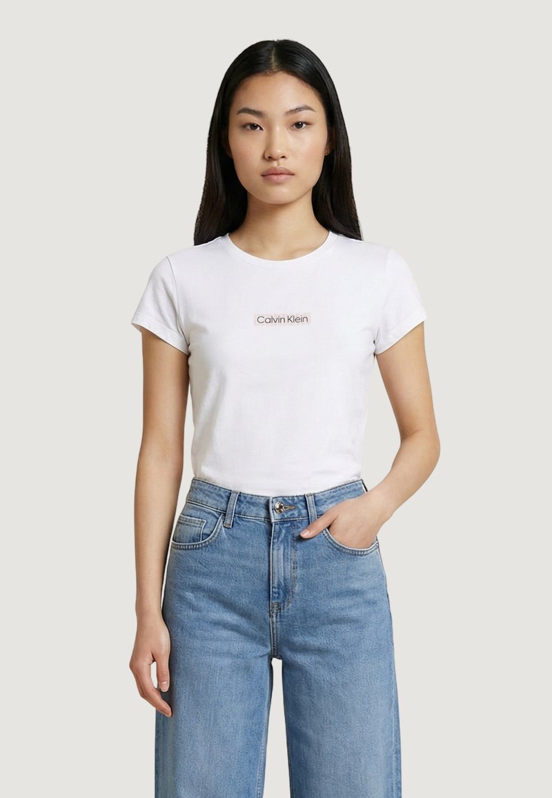 Jeune femme portant un t-shirt blanc Calvin Klein et un jean taille haute bleu clair, debout devant un fond clair uni.
