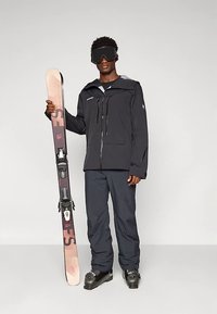 Tamsiai mėlynas slidinėjimo kostiumas, kuriame yra vandeniui atsparus užtraukiamuoju užtrauktuku aprėptas švarkas ir derančios kelnės. Asmuo laiko šviesios spalvos slidę su įjungiamomis tvirtinimo detalėmis.