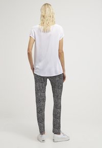 Žena s vlnitými blond vlasy oblečená v bílé krátké tričko, kalhotách s černo-bílým vzorem a bílých teniskách, stojící zády.