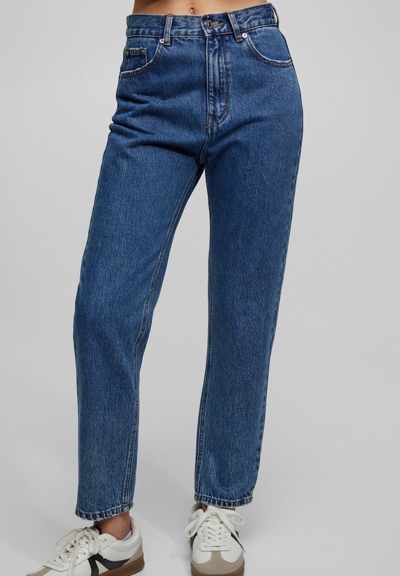 Jean bleu taille haute à jambes droites, porté avec des baskets blanches, montrant de la taille aux chevilles sur un fond gris uni.