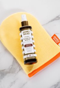 James Read Coconut Glow-Oil Spritz Flasche auf einem gelben und orangefarbenen Bräunungshandschuh, der auf einer Marmoroberfläche platziert ist.